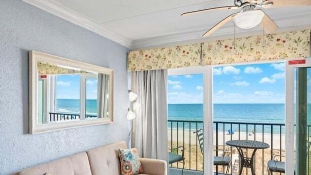 A room with a beige sofa, a small table, and a balcony with a view of the beach and ocean. - Southern Comfort - Expansive Views Of The Ocean And Beach! Newly Renovated Plus Top Grade Linens! 2 Bedroom Condo by RedAwning (Carolina Beach)