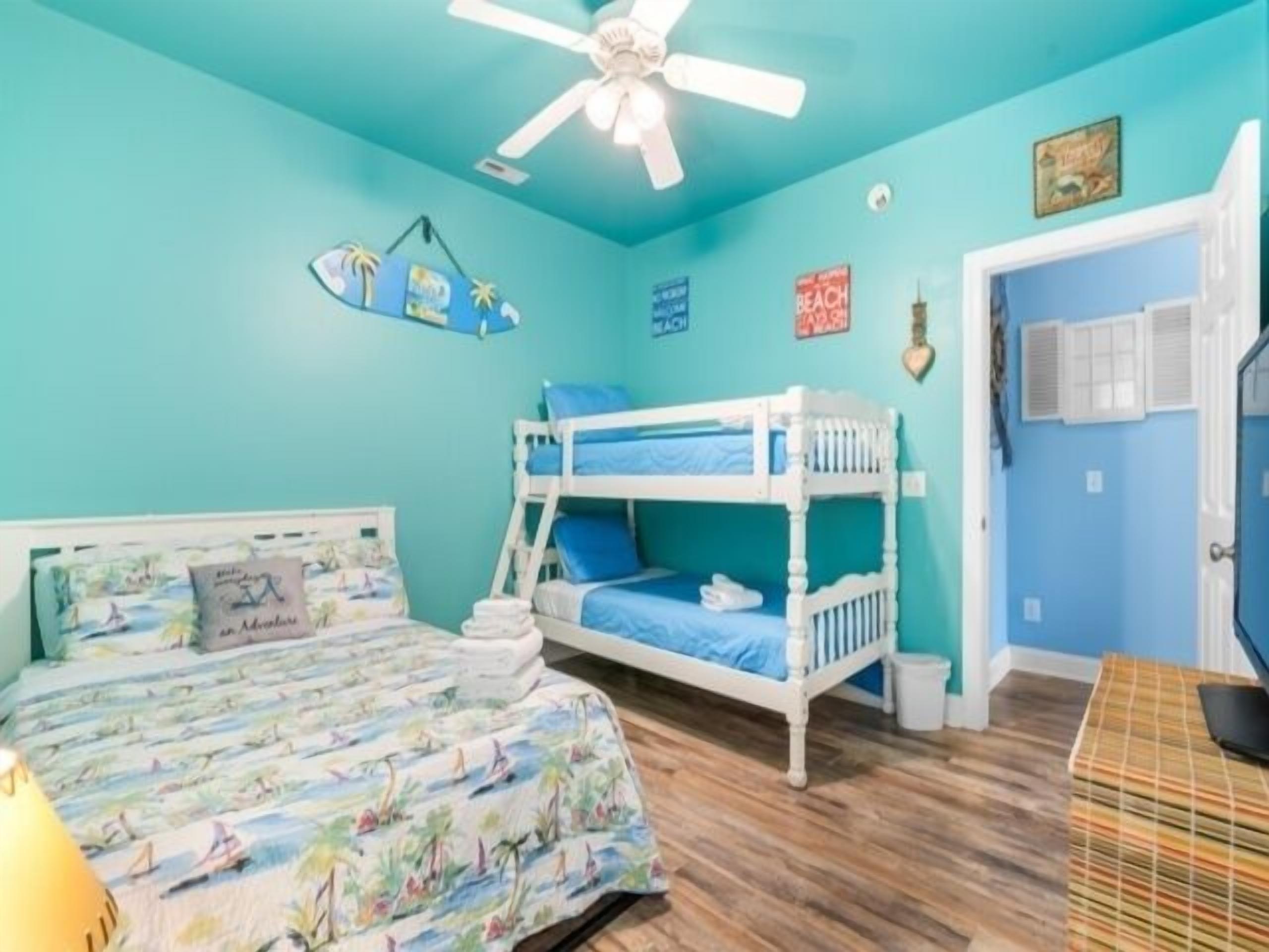 A room with a bunk bed, a single bed with a colorful bedspread, a ceiling fan, and a door with a window.