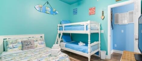 A room with a bunk bed, a single bed with a colorful bedspread, a ceiling fan, and a door with a window.