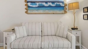 A room with a striped sofa, a wall-mounted painting of a beach scene, a lamp, and framed pictures on the wall. - Life's A Beach - Walk To Everything In Historic Carolina Beach! 1 Bedroom Condo by RedAwning (Carolina Beach)
