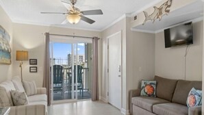 A living room with a ceiling fan, a sofa, a TV, and a sliding glass door leading to an outdoor area. - Life's A Beach - Walk To Everything In Historic Carolina Beach! 1 Bedroom Condo by RedAwning (Carolina Beach)