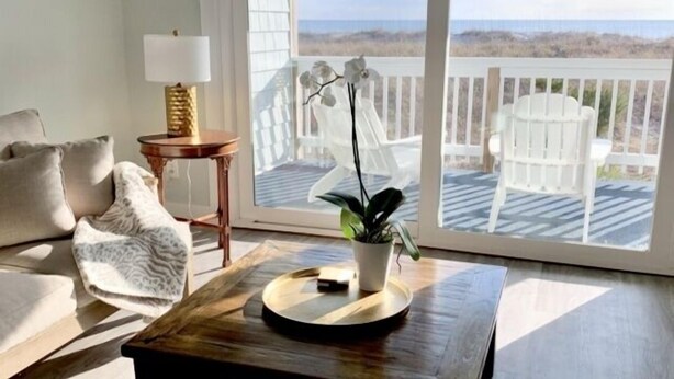 A living room with a view of the beach, a white chair, a wooden coffee table, and a lamp. - Driftwood Cottage - First Floor Oceanfront Beach Retreat! Exceptional Views, Location And Privacy 2 Bedroom Condo by RedAwning (Carolina Beach)