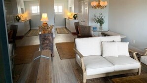 A modern living room with a white sofa, a dining area with a table and chairs, and a chandelier. - Driftwood Cottage - First Floor Oceanfront Beach Retreat! Exceptional Views, Location And Privacy 2 Bedroom Condo by RedAwning (Carolina Beach)