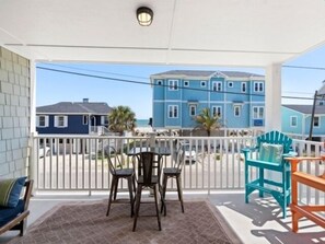 A covered outdoor area with a table and chairs, a white railing, and a view of blue buildings in the background. - Laid Back By The Sea, Steps From The Beach With Amazing Ocean Views by RedAwning (Carolina Beach)