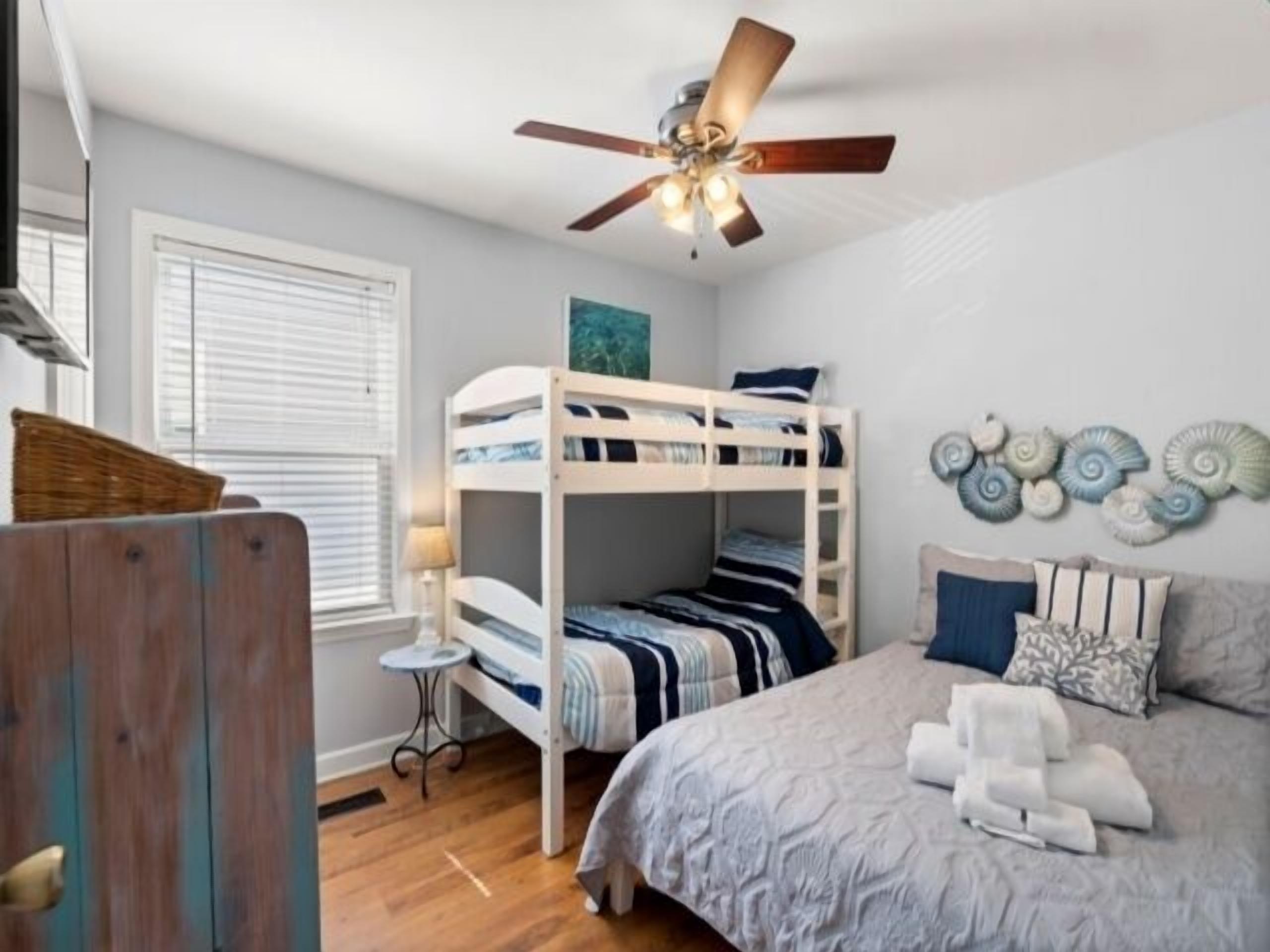 Une chambre avec un lit superposé, un lit simple, un ventilateur de plafond, une petite table et des œuvres d’art décoratives au mur.