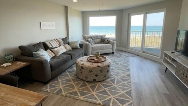 A living room with a sofa, ottoman, and a ceiling fan. There is a large window with a view of the beach. - Carolina Beach Dreamin - Light Filled South Side End Unit. Ocean Views From Most Rooms! Private Beach Access! 3 Bedroom Condo by Redawning (Carolina Beach)