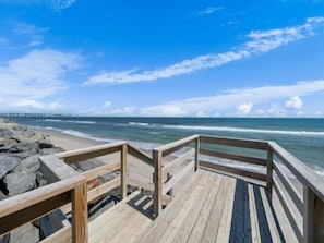 A wooden boardwalk leading to a beach with a pier in the distance.