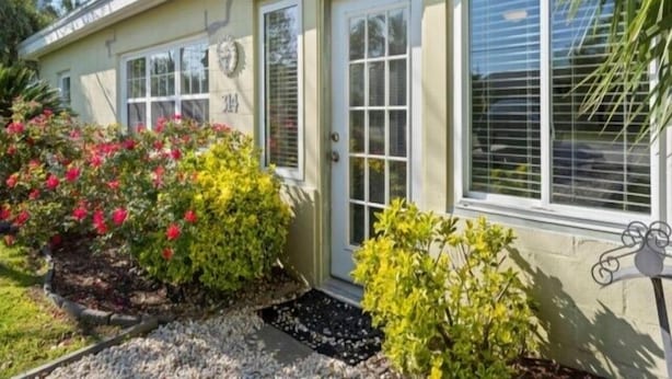 Ein einstöckiges Haus mit einer beigen Fassade, einer Haustür und einer Garagentür. Die Vorderseite hat zwei Fenster und einen kleinen Garten mit Sträuchern und Blumen.