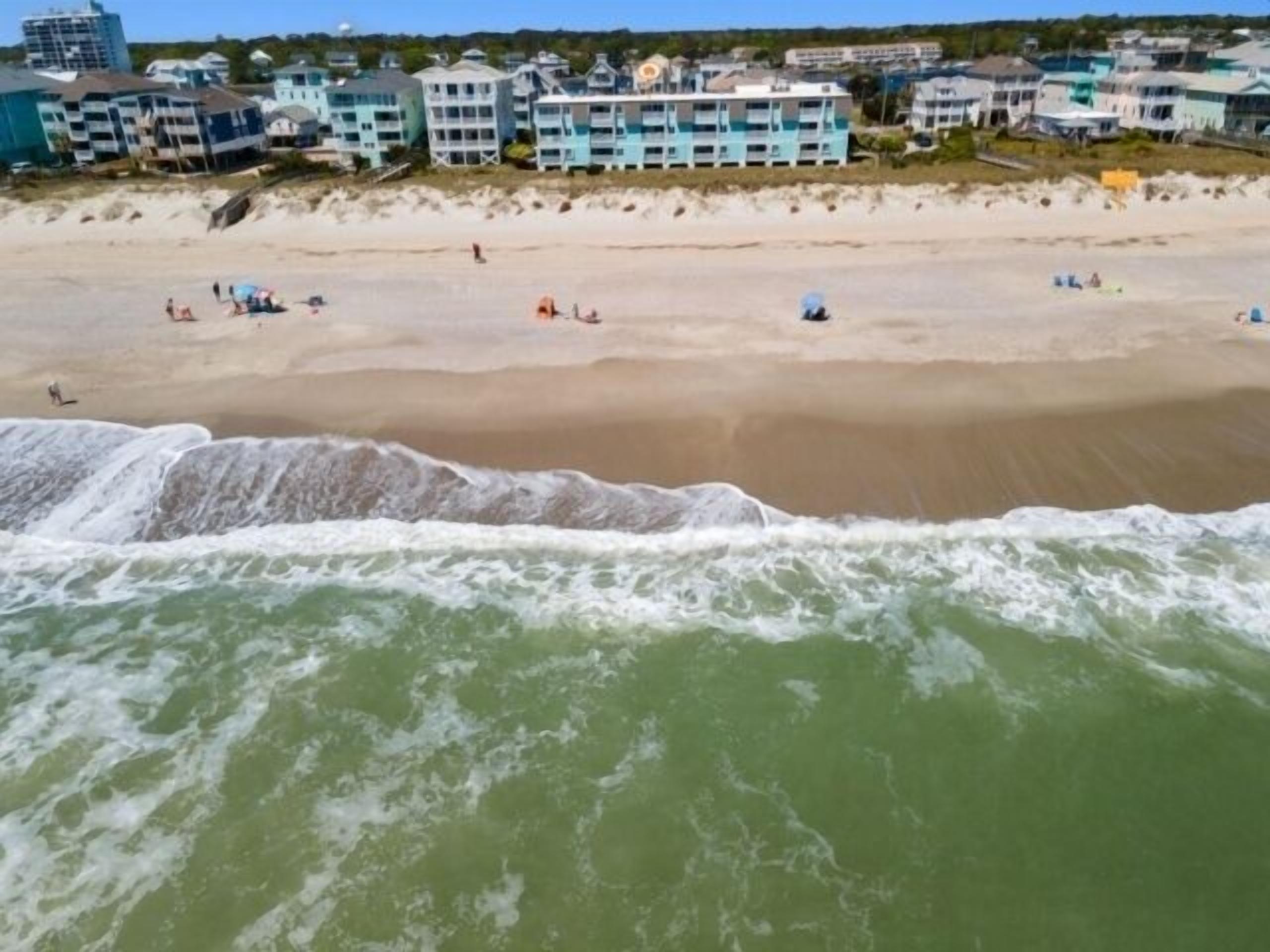 A beach with a wave crashing onto the shore, buildings in the background, and people scattered across the sand.