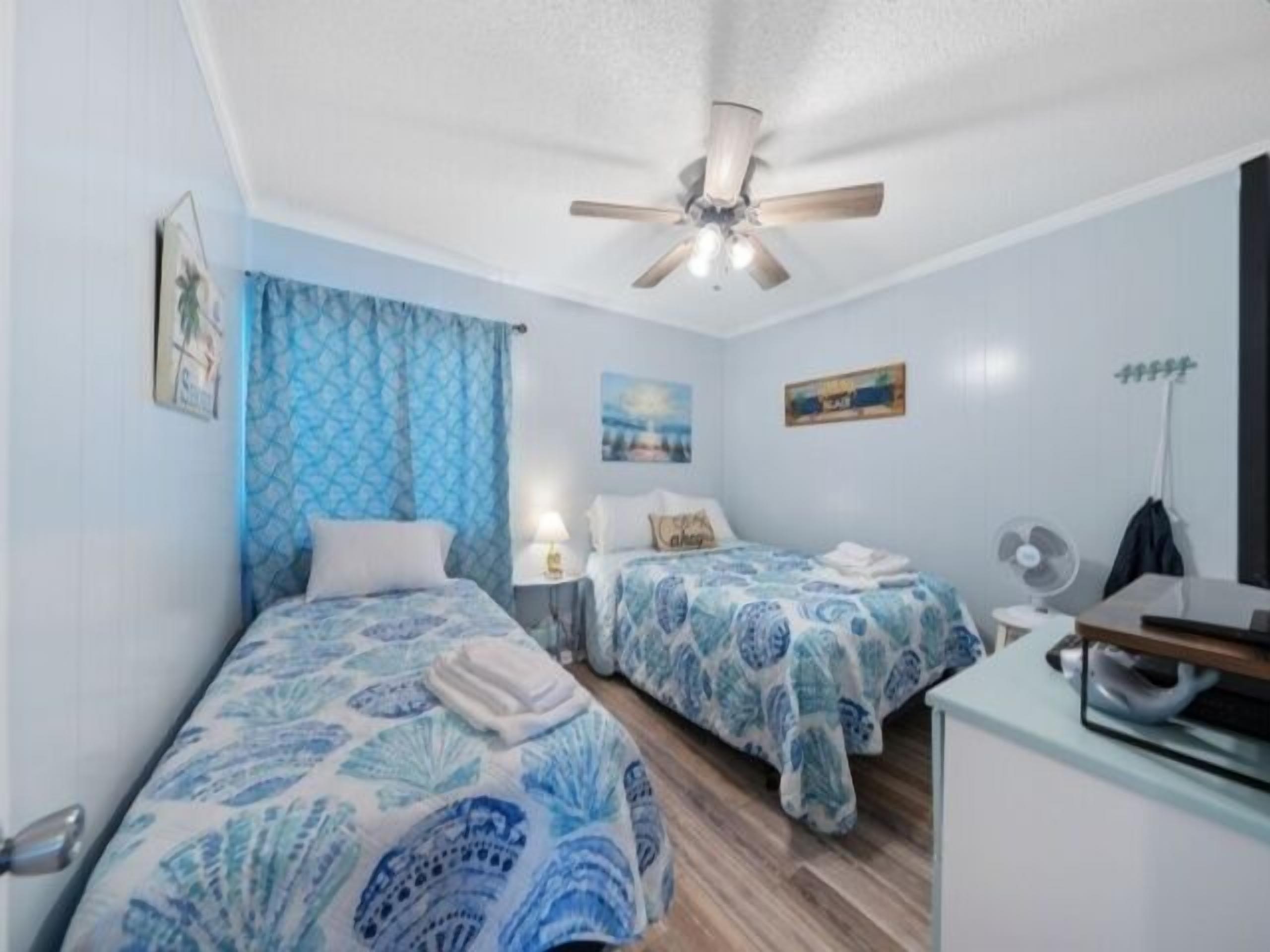 Quarto com duas camas, ventoinha no teto, escrivaninha e ventoinha na parede.