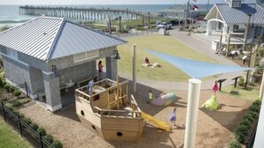 A playground with a slide and climbing structures, a beach view, and a pier in the background. - Sea Haven - Oceanfront! Amazing Master Suite With A Private Oceanfront Deck! Recently Renovated And Perfect For The Entire Family 3 Bedroom Home by RedAwning (Kure Beach)