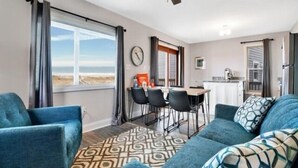A modern living room with a sofa, a dining table with chairs, a ceiling fan, and a view of the outdoors through a window. - Sea Haven - Oceanfront! Amazing Master Suite With A Private Oceanfront Deck! Recently Renovated And Perfect For The Entire Family 3 Bedroom Home by RedAwning (Kure Beach)