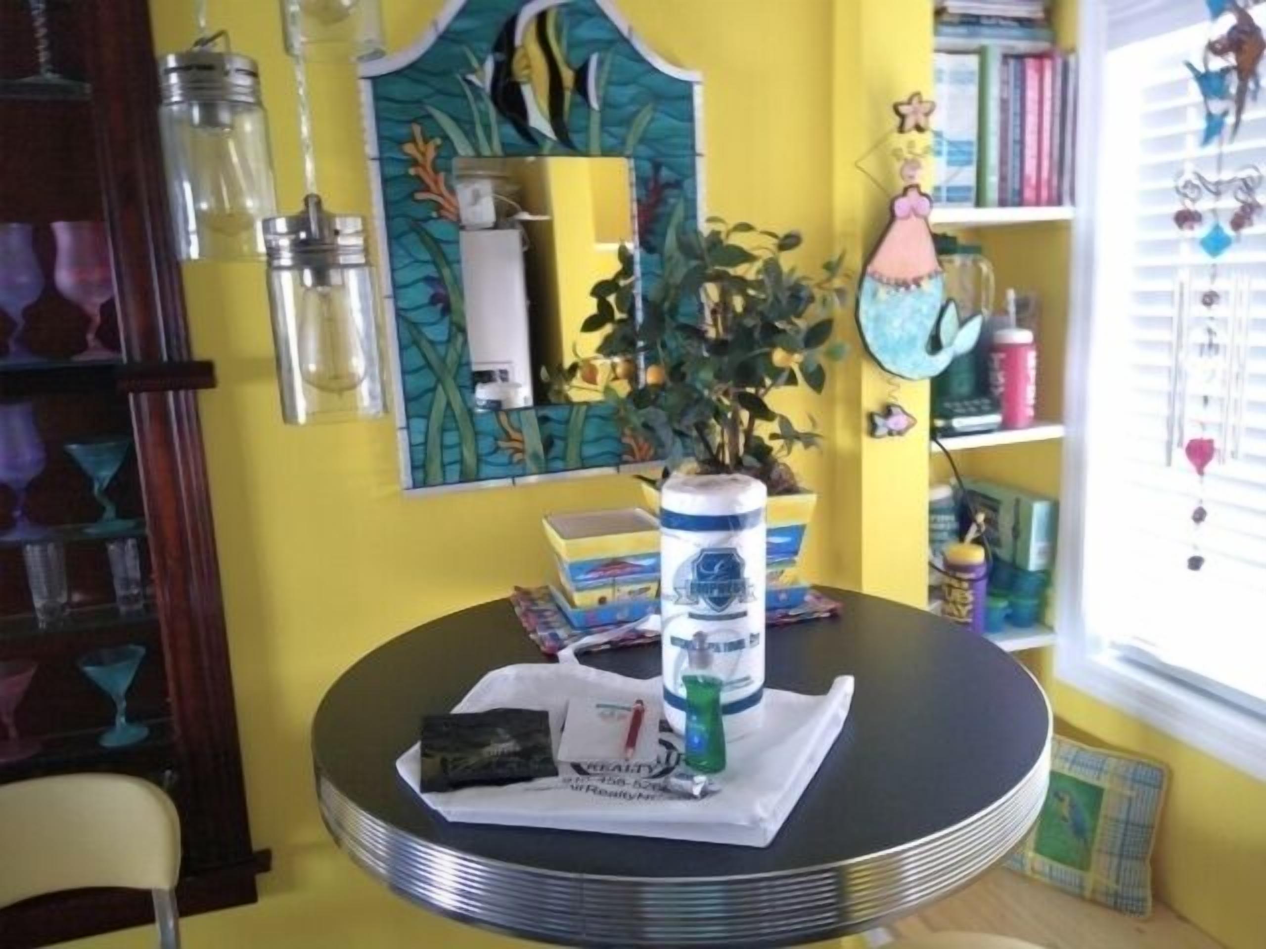 Une table ronde avec une plante, une boîte de conserve et un paquet, dans une pièce aux murs jaunes, avec une bibliothèque et une fenêtre équipée de stores.