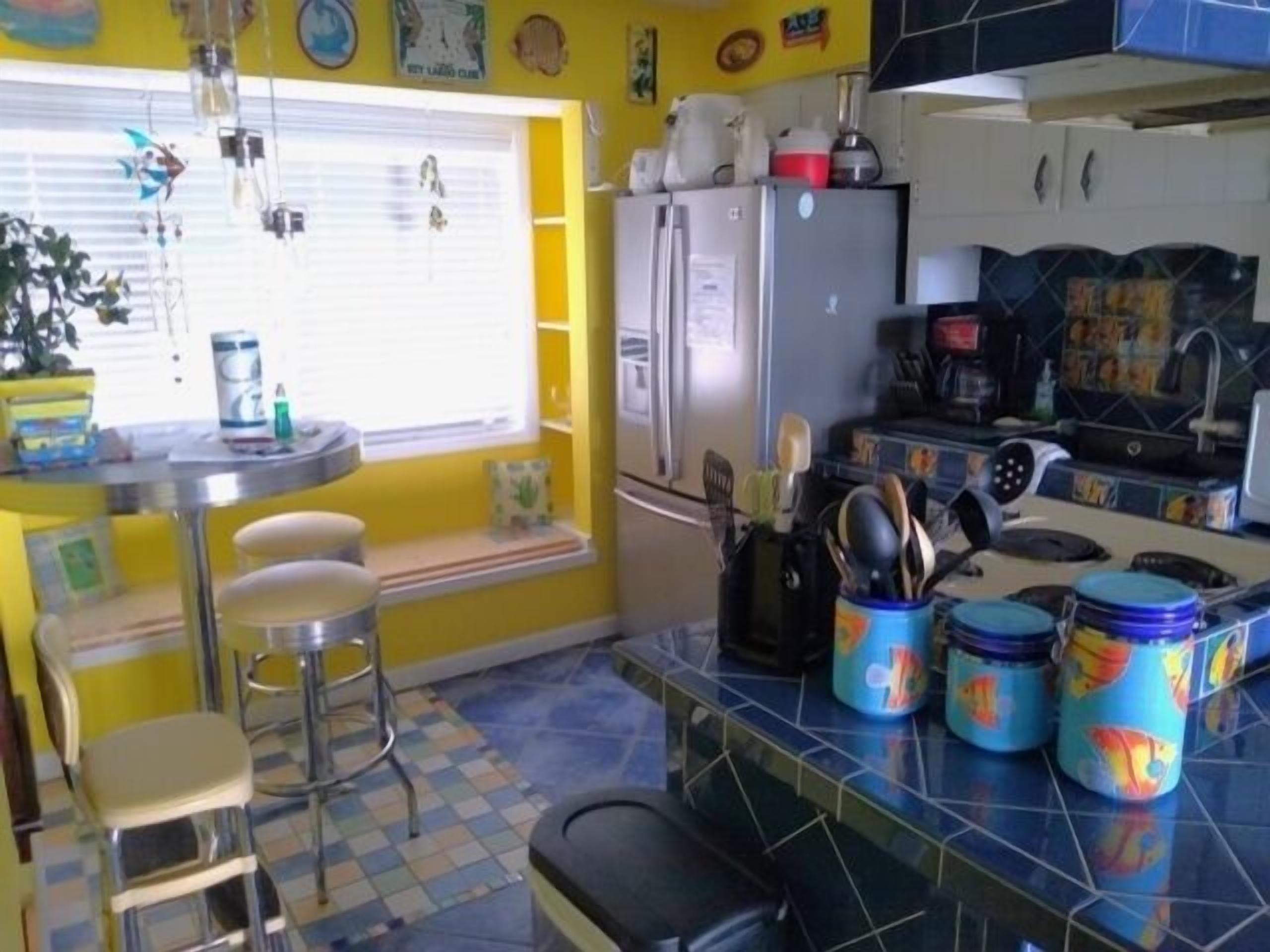 A compact kitchen with a refrigerator, stove, and various kitchen utensils. There is a window with blinds and a small bar area with stools.