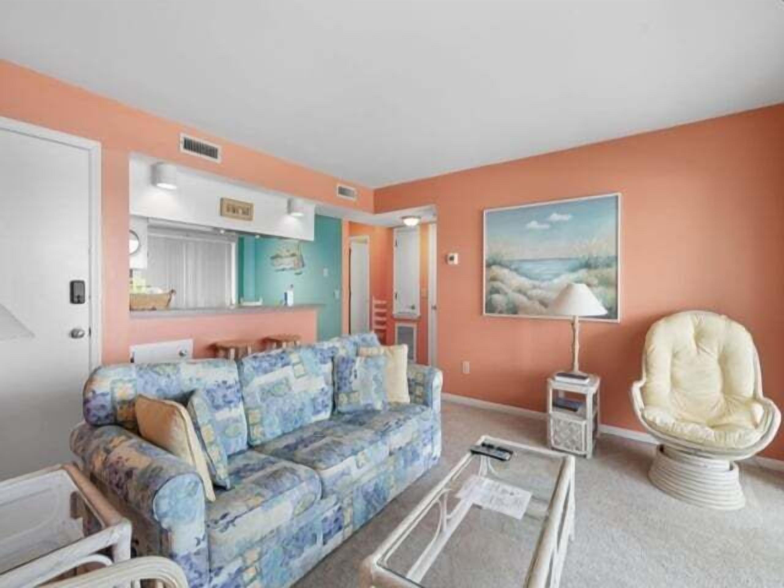 A living room with a floral sofa, a white armchair, a glass coffee table, and a painting of a beach scene.