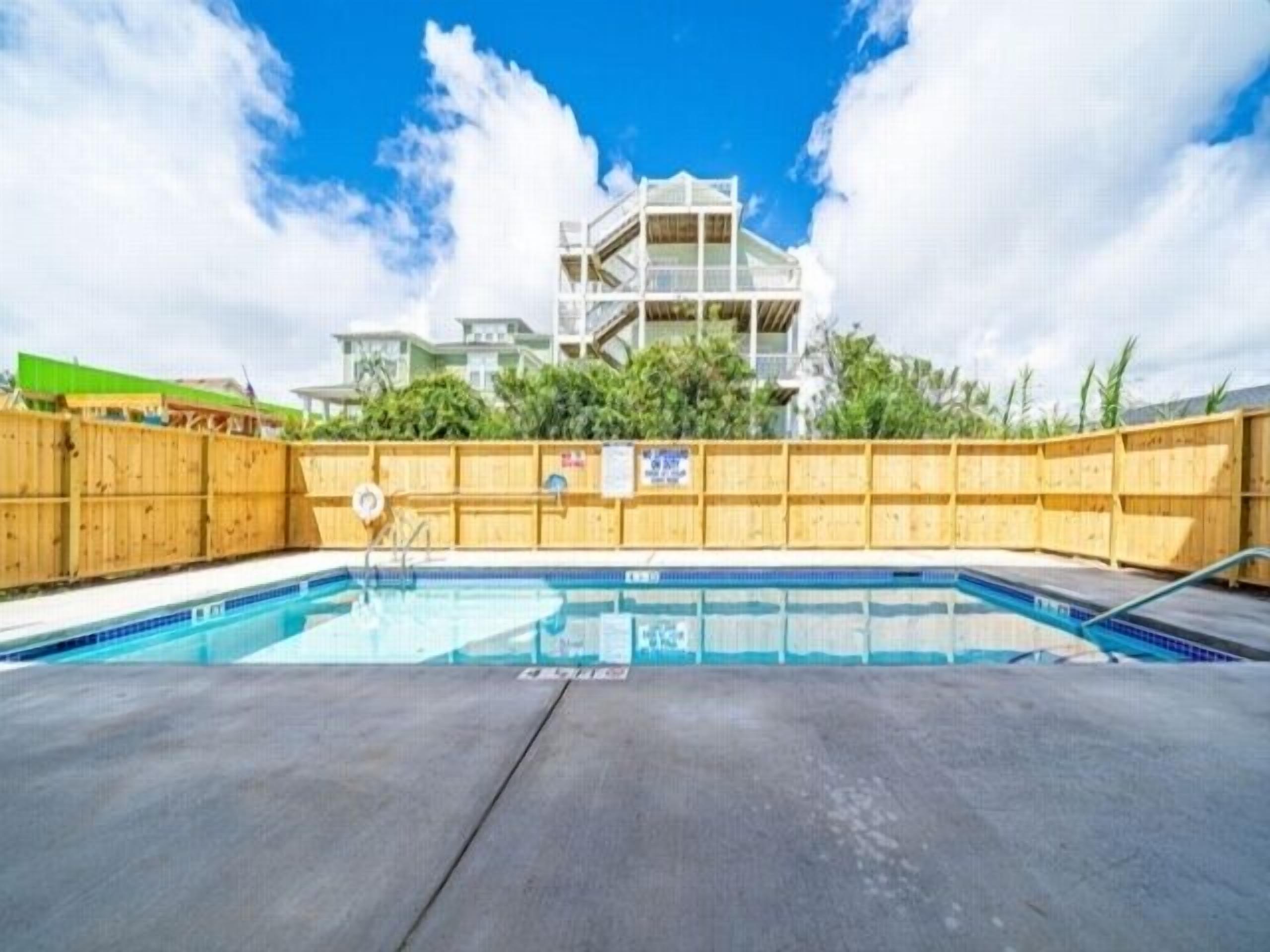 Piscina com cerca de madeira, cadeira de salva-vidas e um edifício com vários andares ao fundo.