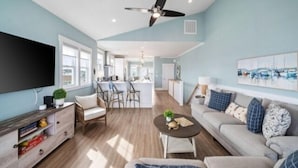 A spacious living room with a large flat-screen TV, a ceiling fan, and a dining area with a kitchen in the background.