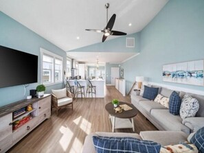 A spacious living room with a large flat-screen TV, a ceiling fan, and a dining area with a kitchen in the background. - Sunnyside - Ocean And Inlet Views, Steps To Beach Access, Plus Parking For 4! 5 Bedroom Townhouse by RedAwning (Carolina Beach)
