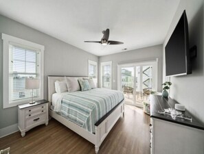 A bedroom with a bed, nightstands, a ceiling fan, a TV, and a view of the outside. - Sunnyside - Ocean And Inlet Views, Steps To Beach Access, Plus Parking For 4! 5 Bedroom Townhouse by RedAwning (Carolina Beach)