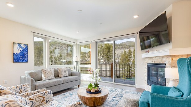 A spacious living room with a grey sofa, teal armchair, and a coffee table with a plant. Large windows offer a view of the outdoors. - 204 Clearwater Lofts 2 Bedroom Condo by RedAwning (Keystone)