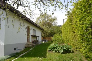 Ein modernes Haus mit heller Fassade, einer überdachten Veranda und einem gepflegten Garten mit verschiedenen Pflanzen und Blumen.