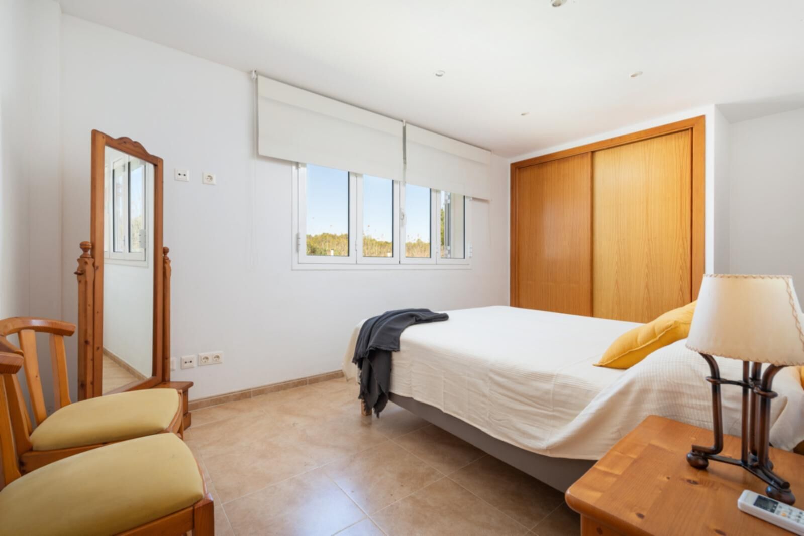 Ein Schlafzimmer mit einem Bett, einem Holzschrank, einem Stuhl, einem Schreibtisch mit Lampe und einem Fenster mit Blick ins Freie.