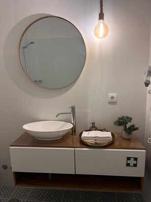 Ett badrum med ett runt spegel, ett fristående handfat, ett träfärgat tvättställsskåp, en liten krukväxt och en lampa.