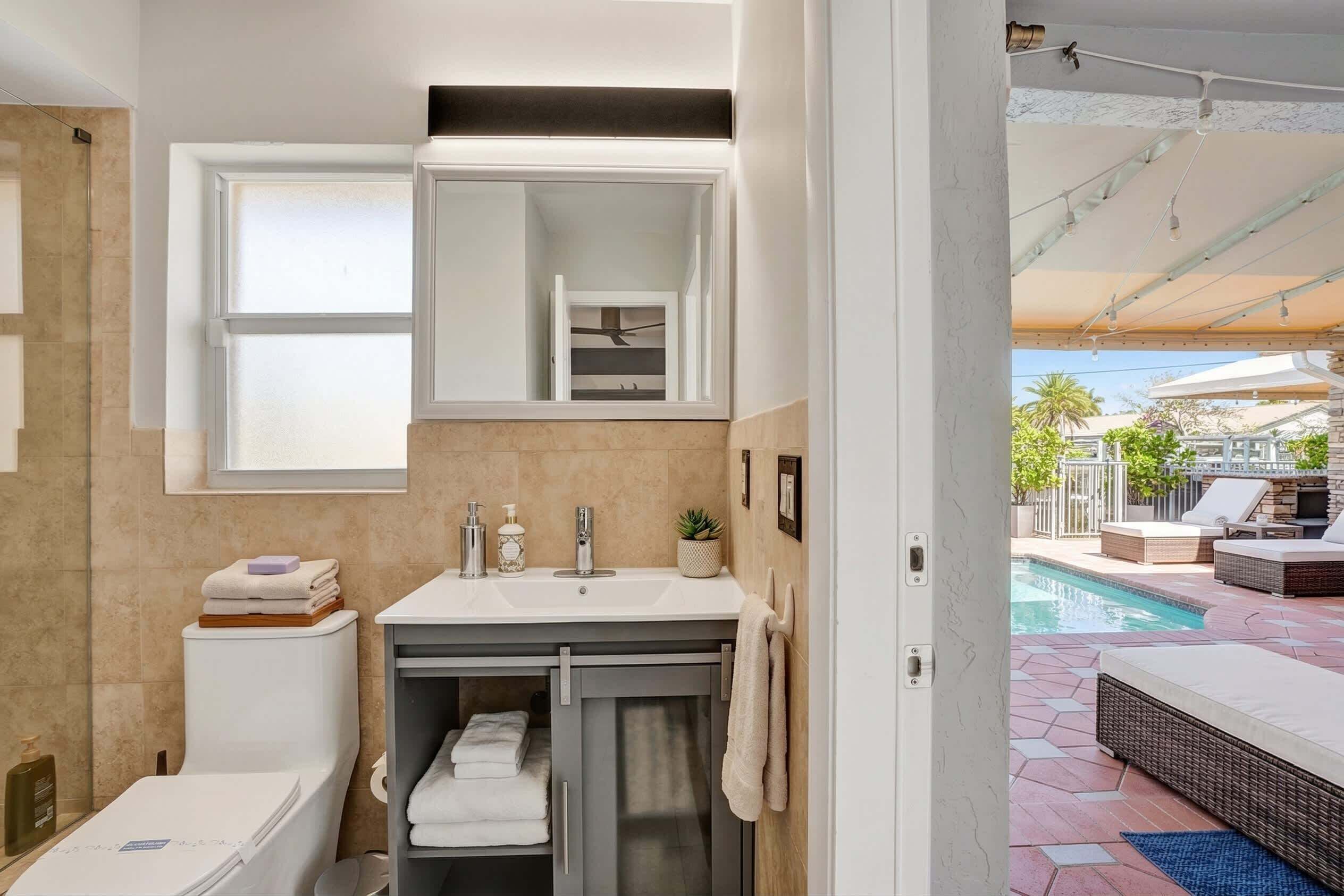A bathroom with a toilet, sink, and mirror, and a view of an outdoor pool area.