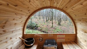 Eine Holzhütte mit einem großen Bogenfenster mit Blick auf einen Wald. Im Inneren befinden sich eine hölzerne Bank, ein Metallgestell und ein schwarzer Topf mit einem Holzgriff.