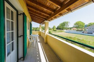 Un balcón con techo de madera, sillas blancas y vista a un área verde y edificios al fondo.