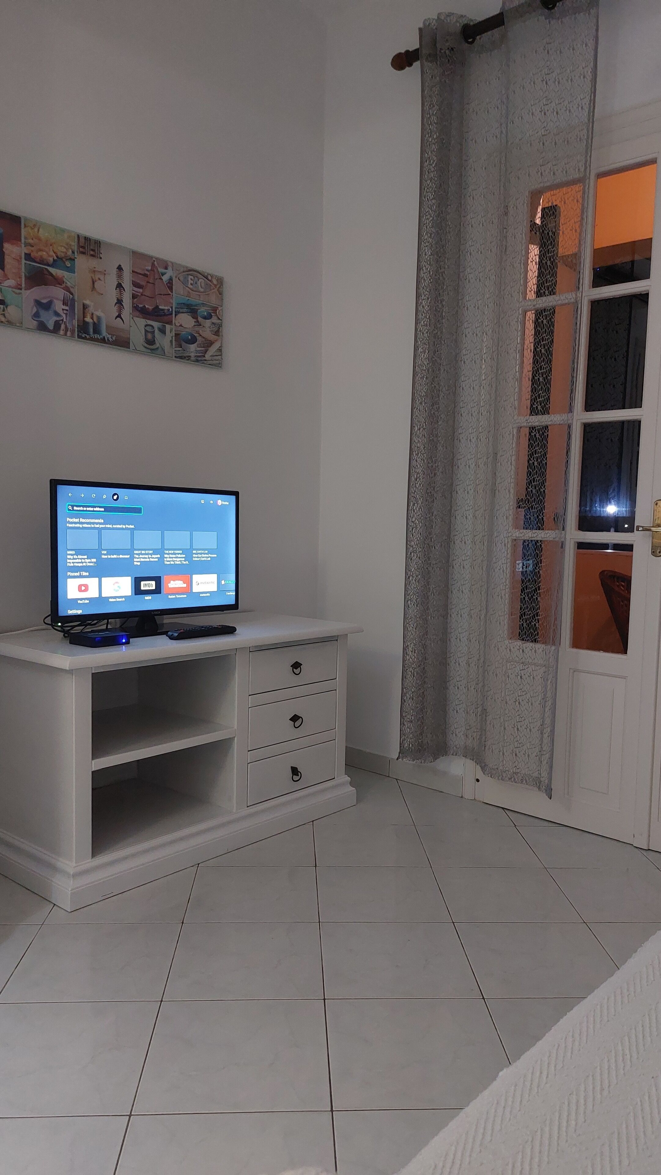 Una habitación con un televisor blanco sobre un mueble, un pequeño armario blanco con cajones, piso de baldosas y una puerta con mosquitera.