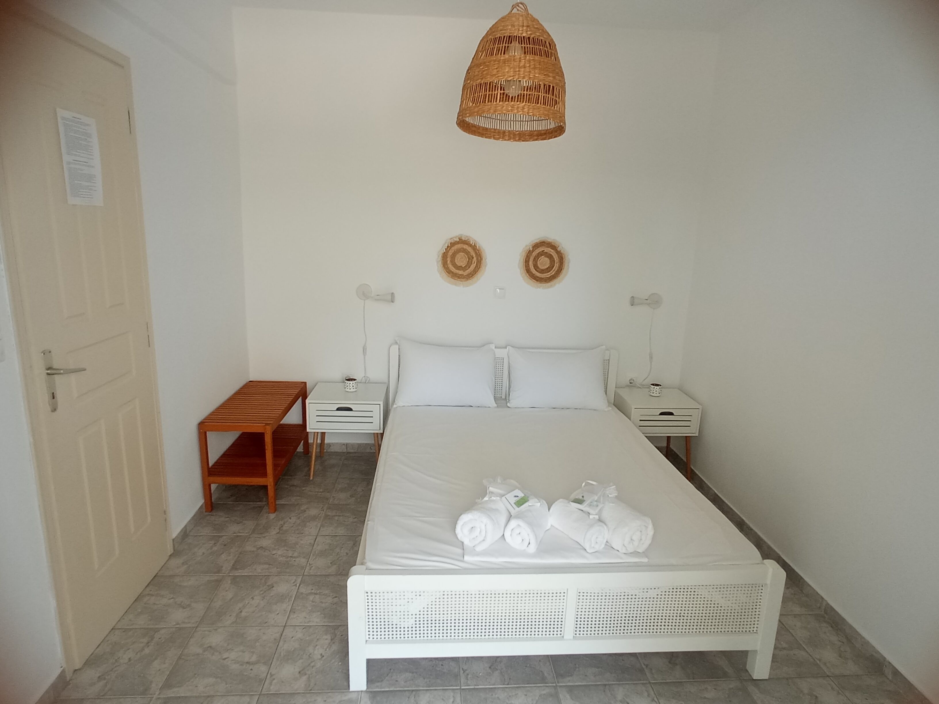 A bedroom with a bed, bedside tables, a wooden bench, a woven light fixture, and two wall decorations.