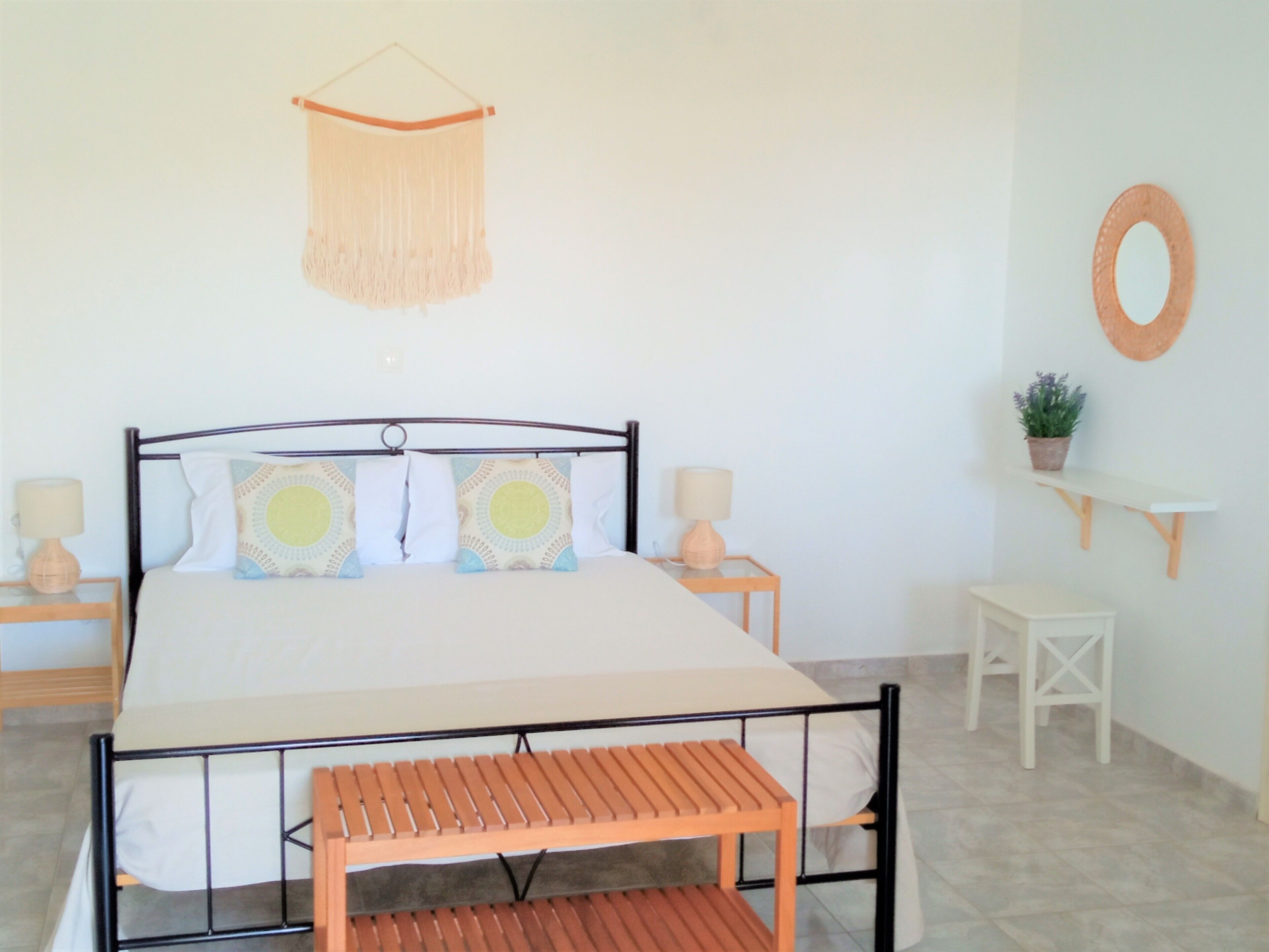 A bedroom with a bed, bedside tables, a hanging macrame, a small shelf with a plant, and a round mirror on the wall.