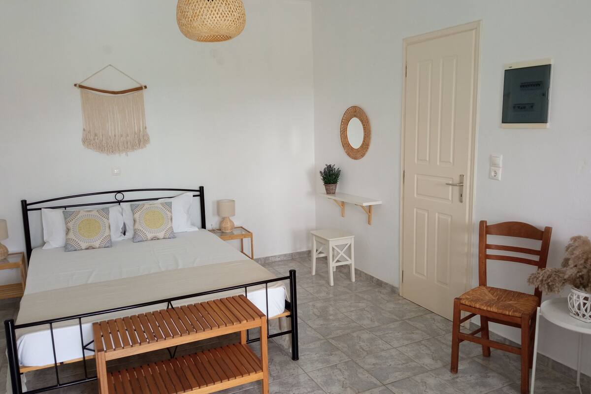 A bedroom with a bed, bedside tables, a bench, a chair, a hanging light, a wall-mounted shelf, and a door.