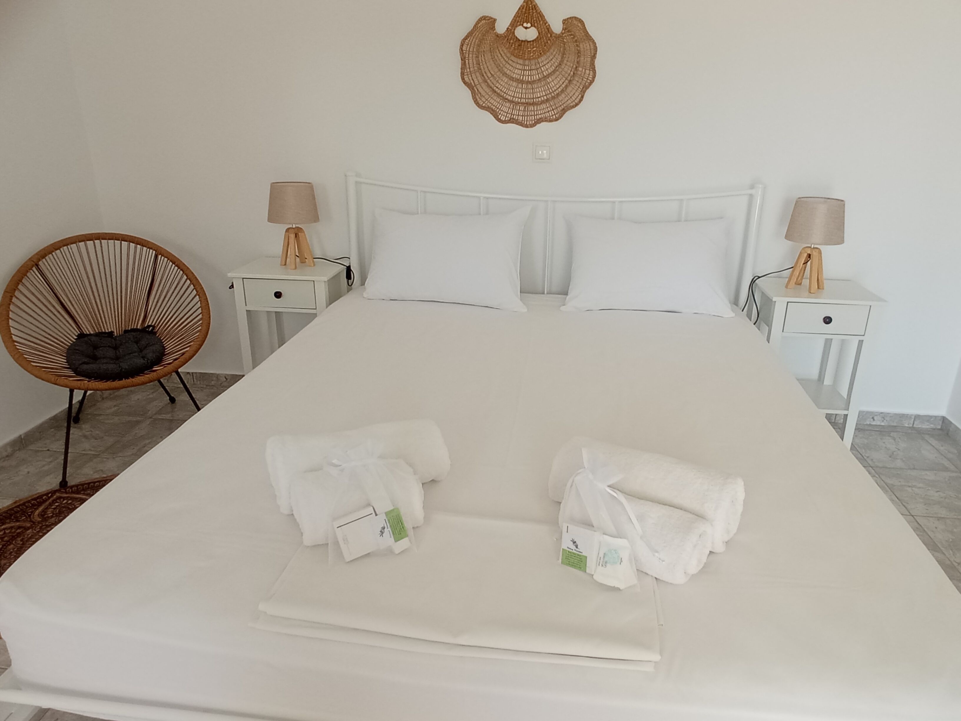A neatly made bed with two towels, a wicker chair, and two bedside tables with lamps.