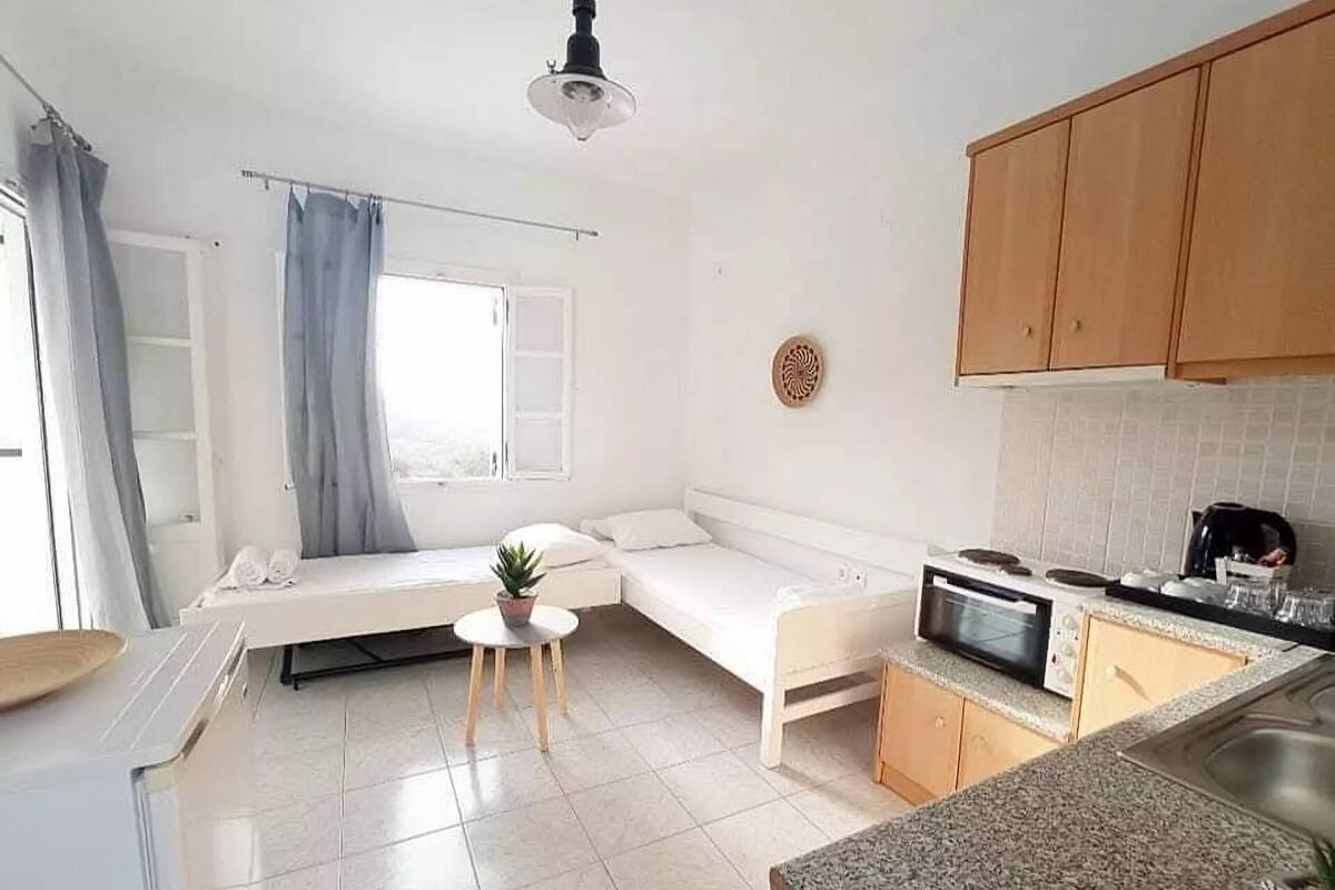 A compact kitchen with a microwave, stove, and sink. There is a small dining table with a plant. A bed with a window and curtains is visible.