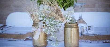 Two rustic jars with flowers and wheat, placed on a table with lace and a brick wall in the background.