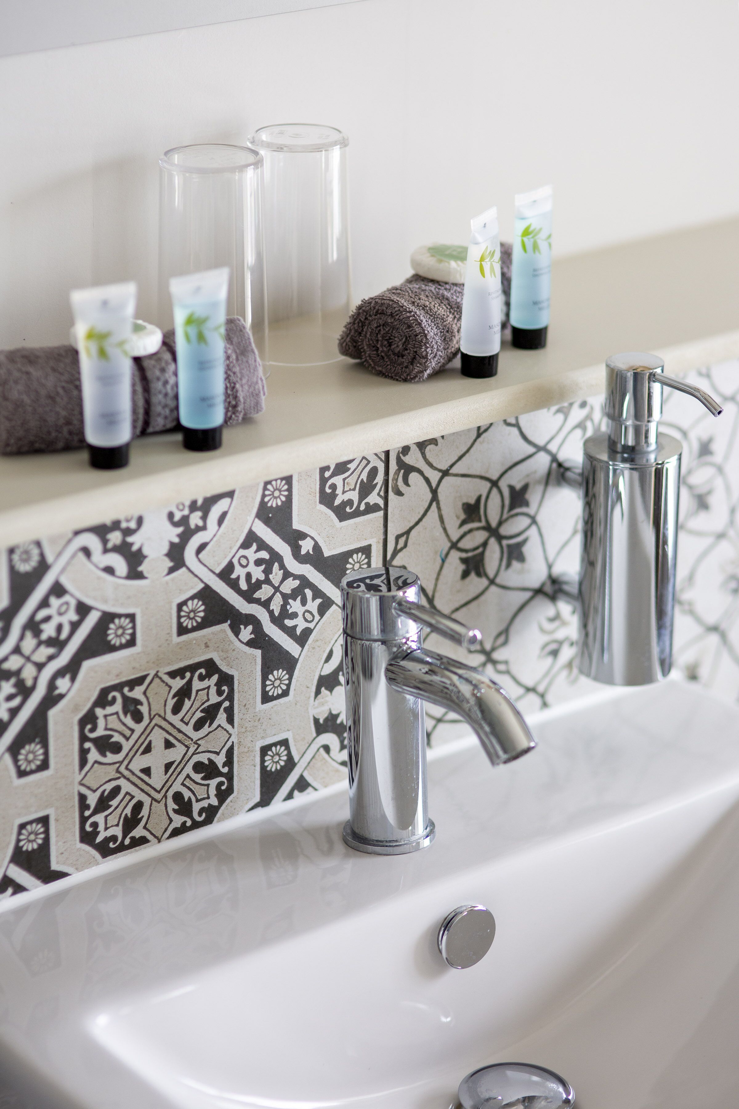 A bathroom with a white sink, two chrome faucets, and a patterned backsplash.