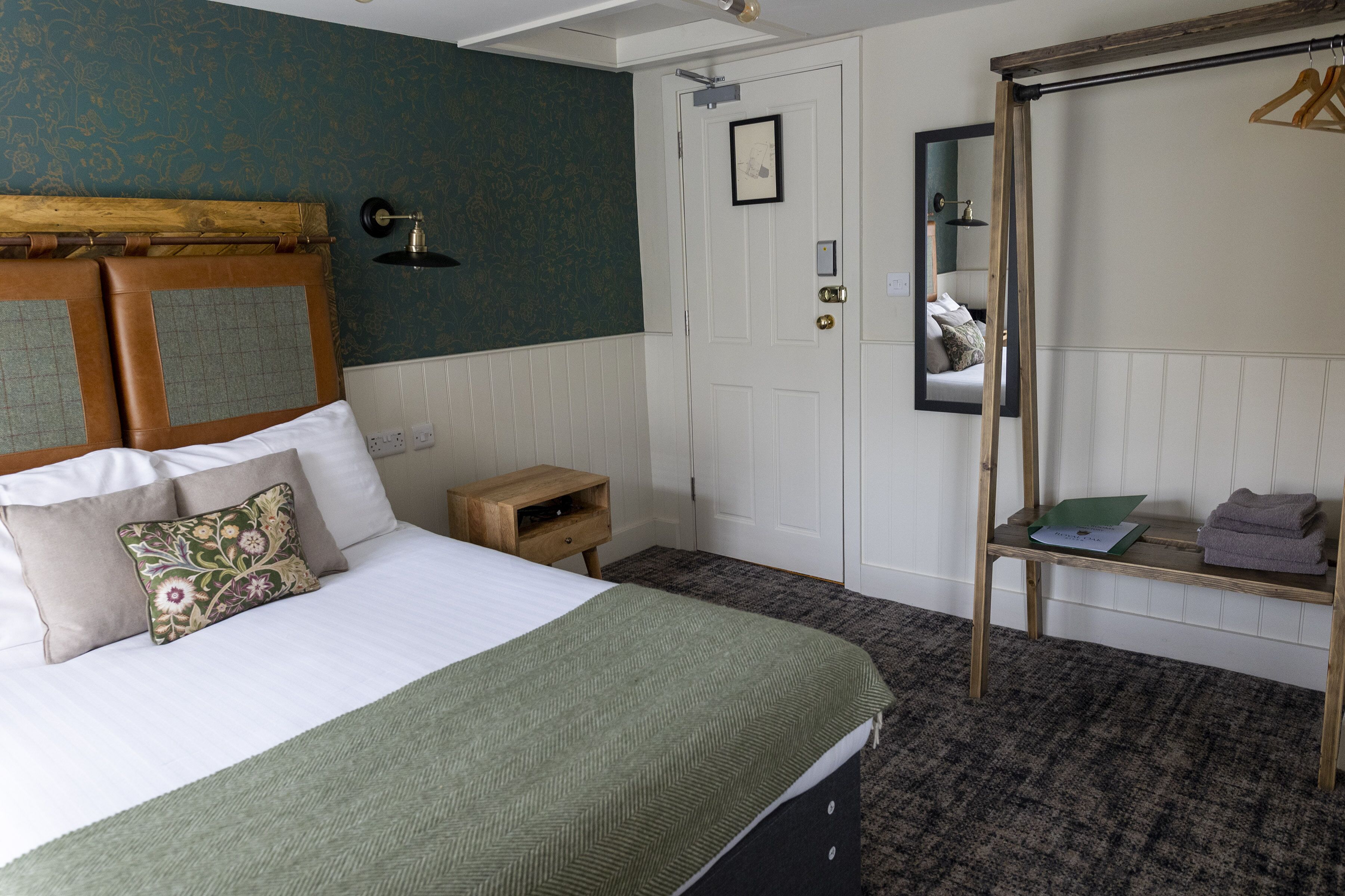 A neatly arranged bedroom with a bed, a wooden headboard, a small bedside table, a mirror, and a clothing rack.