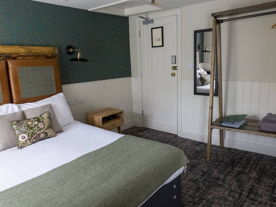 A neatly arranged bedroom with a bed, a wooden headboard, a small bedside table, a mirror, and a clothing rack.