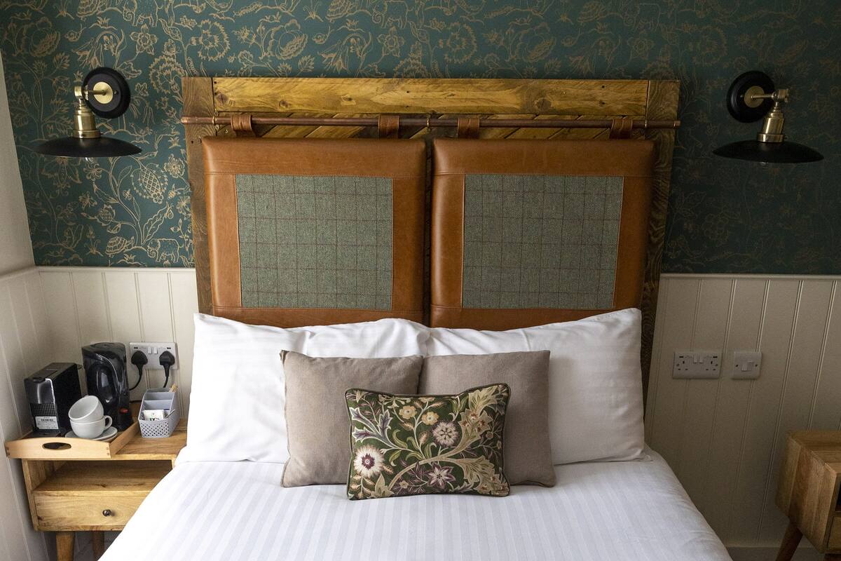 A neatly made bed with a wooden headboard, a bedside table with a kettle and cups, and a patterned wall.