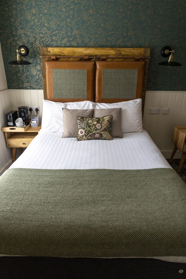 A neatly made bed with a wooden headboard, a bedside table with a kettle and cups, and a patterned wall. - Royal Oak (Dover)