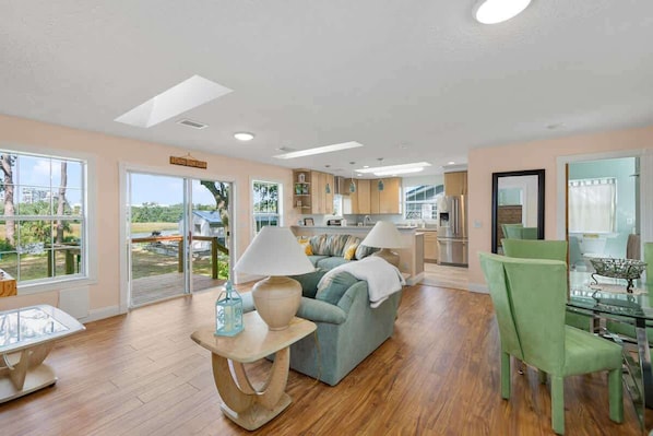 A spacious living area with a sofa, a dining table with chairs, a kitchen in the background, and large windows offering a view of the outdoors.