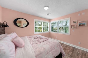 A bedroom with a bed, a round mirror, two windows with blinds, and a ceiling light.