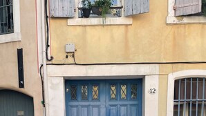 Een blauwe deur met daarboven het nummer '12', een raam met tralies en een klein balkon met planten.
