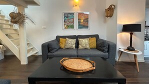 Een woonkamer met een grijze bank, twee ingelijste foto’s, een zwarte salontafel met een gevlochten dienblad, een lamp en een trap.