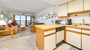 A spacious living area with a large window overlooking the beach, a kitchen with wooden cabinets, and a dining area with wicker furniture.