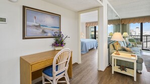 Ein Hotelzimmer mit Holztisch, einem Sessel, einer Couch, einem kleinen Tisch mit Lampe, einem Gemälde von einem Leuchtturm und Meerblick durch große Fenster.