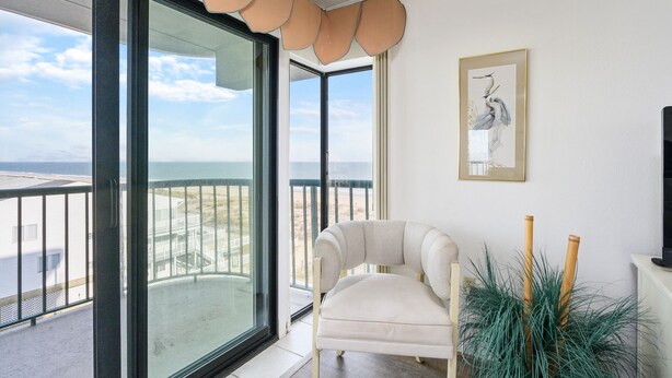 Ein Balkon mit Blick auf Strand und Meer, ein weißer Sessel, ein gerahmtes Kunstwerk und ein Blumentopf.
