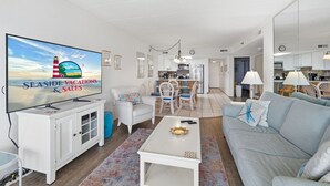 Ein Wohnzimmer mit Fernseher, der 'Seaside Vacations & Sales' zeigt, einer Couch, einem Couchtisch und einem Essbereich mit Stühlen und Tisch.